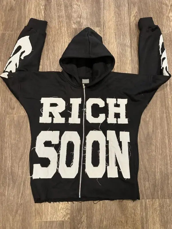 Front view of black zip-up hoodie featuring 'RICH SOON' text