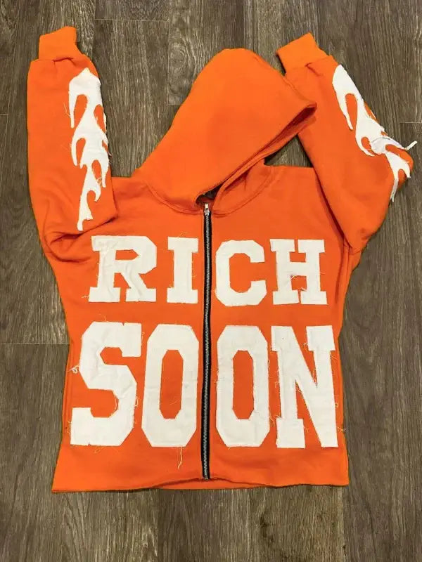 Front view of orange zip-up hoodie featuring 'RICH SOON' text