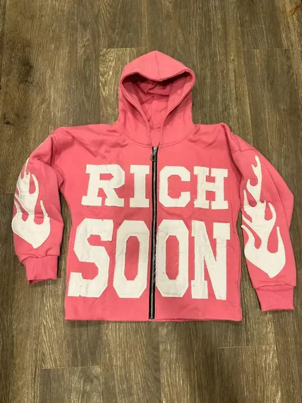 Front view of pink zip-up hoodie featuring 'RICH SOON' text