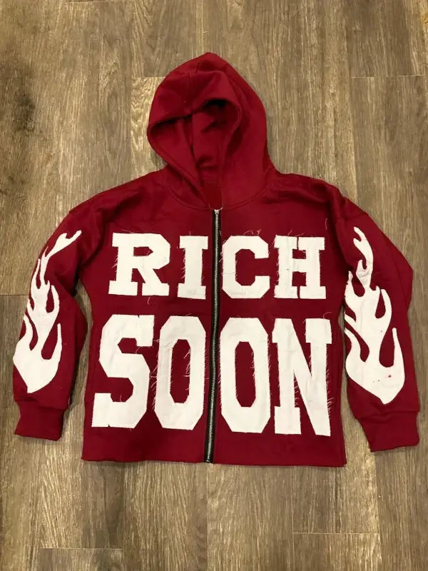 Front view of red zip-up hoodie featuring 'RICH SOON' text 